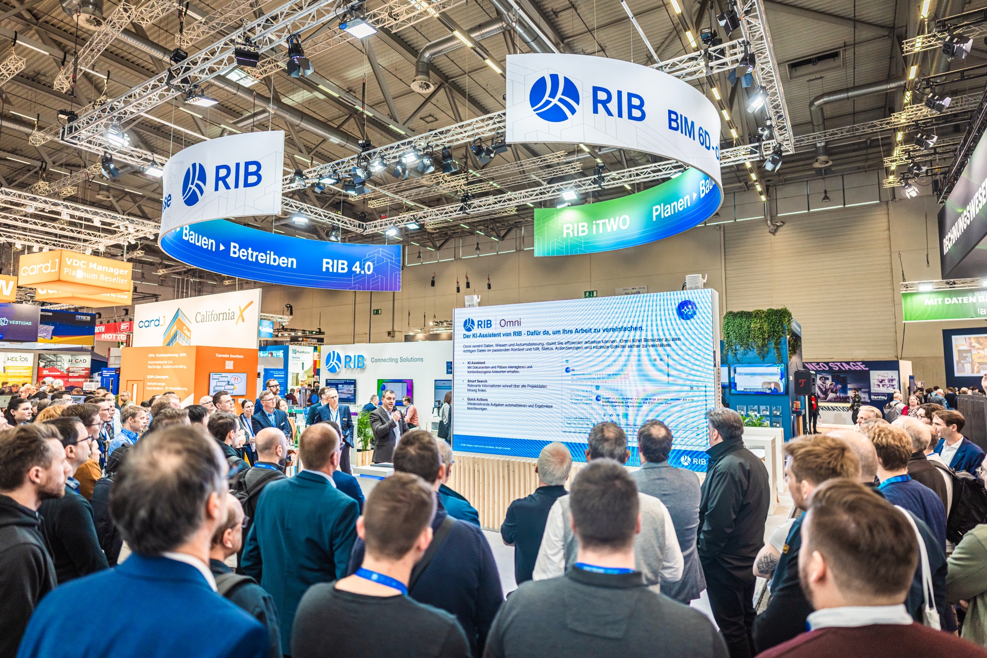 Besucherinnen und Besucher verfolgen eine Produktpräsentation am Messestand von RIB Software auf der BAU in München